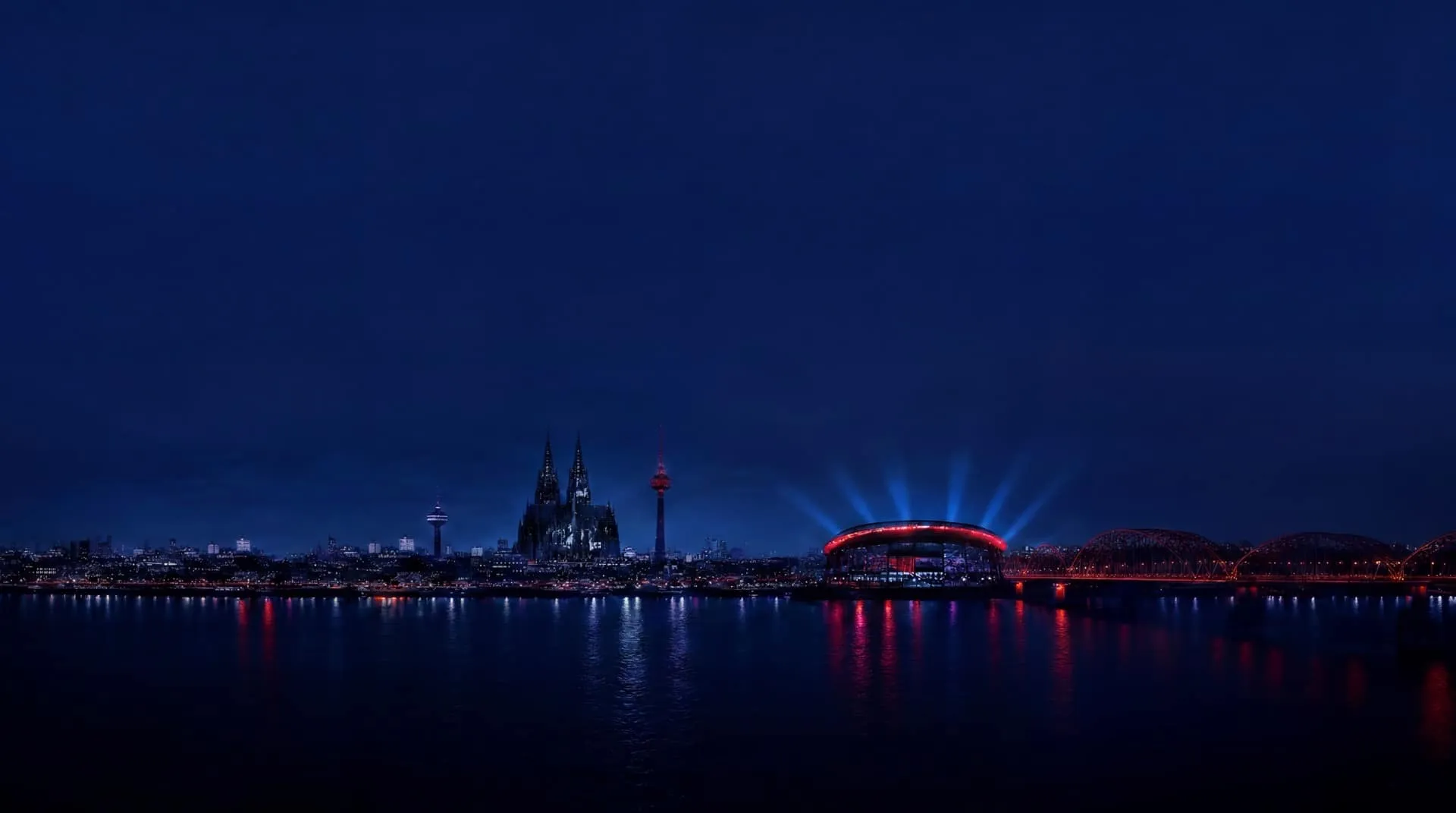 Köln bei Nacht über dem Rhein: Dom, Fernsehturm und die angestrahlte Arena.